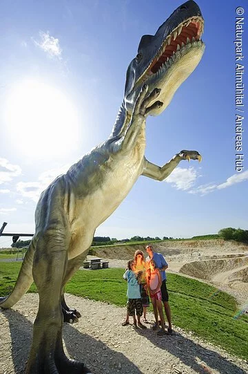 Eine Familie steht am Eingang des Steinbruchs und bestaunt den lebensgroßen Dinosaurier. Im Hintergrund ist der Steinbruch zu sehen.