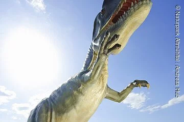 Eine Familie steht am Eingang des Steinbruchs und bestaunt den lebensgroßen Dinosaurier. Im Hintergrund ist der Steinbruch zu sehen.