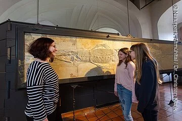 Vor der riesigen Versteinerung eines Fisches, der senkrecht an der Wand hängt, steht links eine Frau im gestreiften Pullover. Sie redet mit zwei Mädchen, die zwei Meter von ihr entfernt stehen.