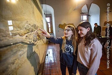 Zwei Mädchen, eine blond und die andere braunhaarig stehen vor dem großen versteinertem Fisch der senkrecht im Raum steht. Im Hintergrund sind weitere Besucher zu erkennen.