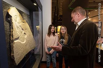 Zwei Mädchen, eine blond und die andere braunhaarig stehen neben einer Versteinerung und schauen interessiert zu dem Mann mit braunem Jackett, der ihnen mehr zu den Fossilien erzählt.