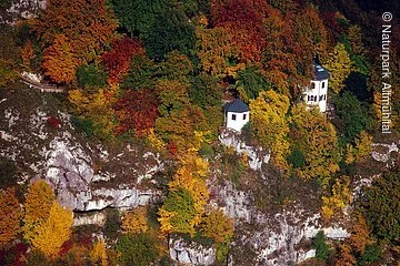 Das Gebiet der Tropfsteinhöhle von oben fotografiert. Die zwei Gebäude blitzen durch das bunte Herbstkleid des Waldes durch.
