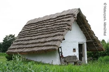 Es ist ein originalgetreue, begehbare Rekonstruktion eines Bajuwarenhauses zu sehen. Es hat ein Strohdach und verkalkte, weiße Außenmauern.