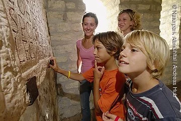 Kinder entdecken das Römerkastell Pfünz Zwei Jungen betrachten im Römerkastell intensiv eine Steintafel mit einer Inschrift. Hinter ihnen stehen zwei lächelnde Frauen, die ihren Blick ebenfalls auf die Inschrift richten.