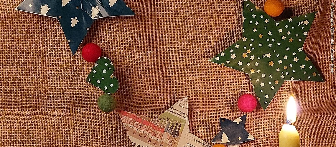 Weihnachtsgirlande mit Papiersternen und Filzkugeln, davor eine brennende Kerze