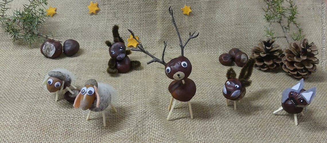 Verschiedende gebastelte Kastanientiere stehen weihnachtlich dekoriert nebeneinander. Links zwei Schafe, daneben folgen ein Eichhörnchen, ein Hirsch, ein Fuchs und ganz rechts ein Esel.