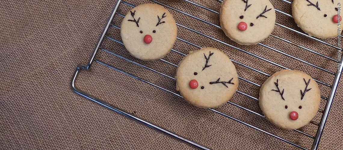 Rudolf Plätzchen liegen auf einem Ofengitter (Helle, runde Butterplätzchen mit roter Smartie-Nase und Schokoladen-Augen und -Geweih). Fünf Plätzchen sind dekoriert und zwei noch nicht.