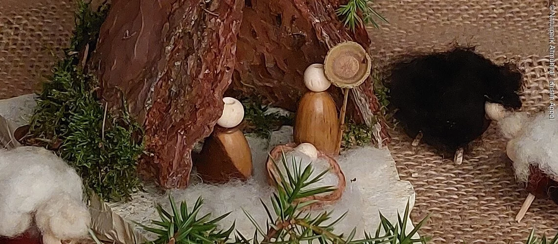 Auf einer weihnachtlich dekorierter Decke steht eine kleine Krippe aus Rinde und Naturstein. Die Krippenfiguren sind aus Eicheln gestaltet.