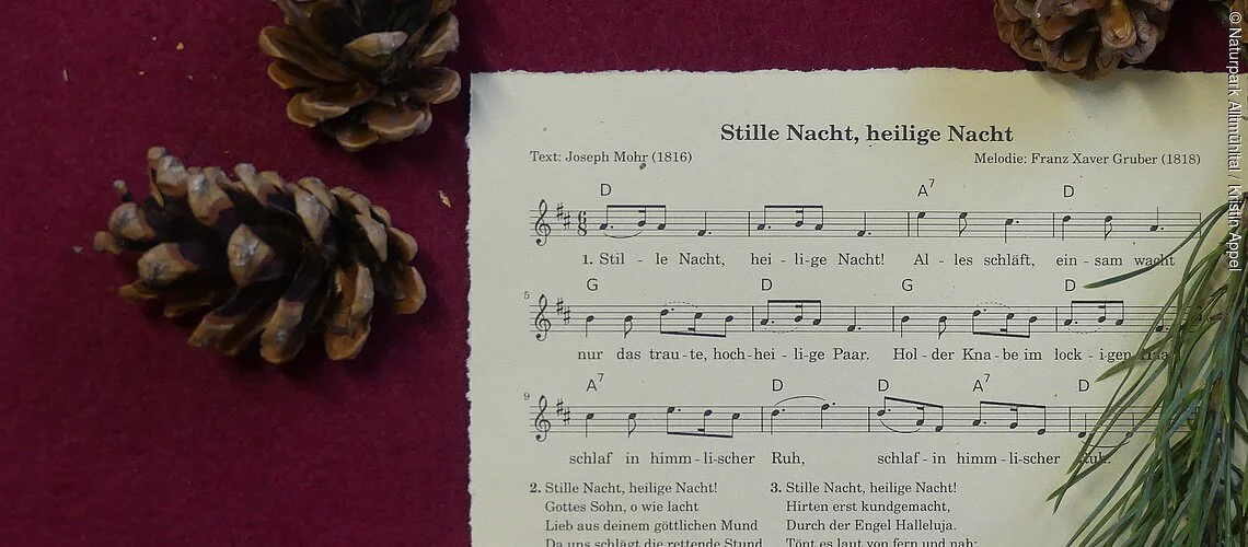Stille Nacht, heilige Nacht Das Notenblatt Stille Nacht, heilige Nacht liegt auf einem weihnachtlich dekorierten Tisch mit Kerze, Tannengrün und Zapfen