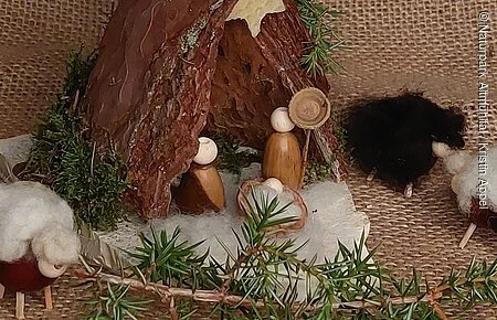 Auf einer weihnachtlich dekorierter Decke steht eine kleine Krippe aus Rinde und Naturstein. Die Krippenfiguren sind aus Eicheln gestaltet.
