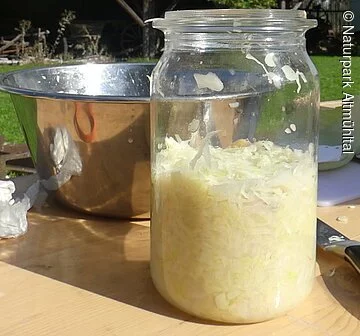 Wir sehen ein Weckglas, das zur Hälfte gefüllt ist mit fermentierten Sauerkraut. Im Hintergrund sieht man den herbstlichen Bauerngarten des Jura-Bauernmhof-Museums mit bewölktem Himmel