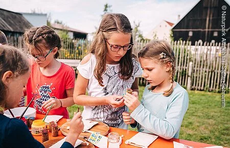 Kinderveranstaltung im Jura-Bauernhof-Museum Vier Mädchen stehen an einem Biertisch im Garten des Jura-Bauernhof-Museums und basteln gemeinsam beim museumpädagischen Programm "ganz schön beeinDRUCKend".