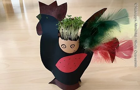 In einem aus Papier gebastelten Hahn steht eine mit Gesicht versehene Eierschale. Aus dem Ei wächst Kresse heraus.