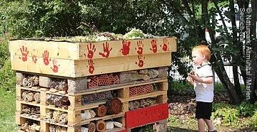 Wildbienenhotel Ein kleiner Junge begutachtet das selbstgebaute Wildbienenhotel, auf dem die Handabdrücke der kleinen Helfer in rot zu sehen sind.