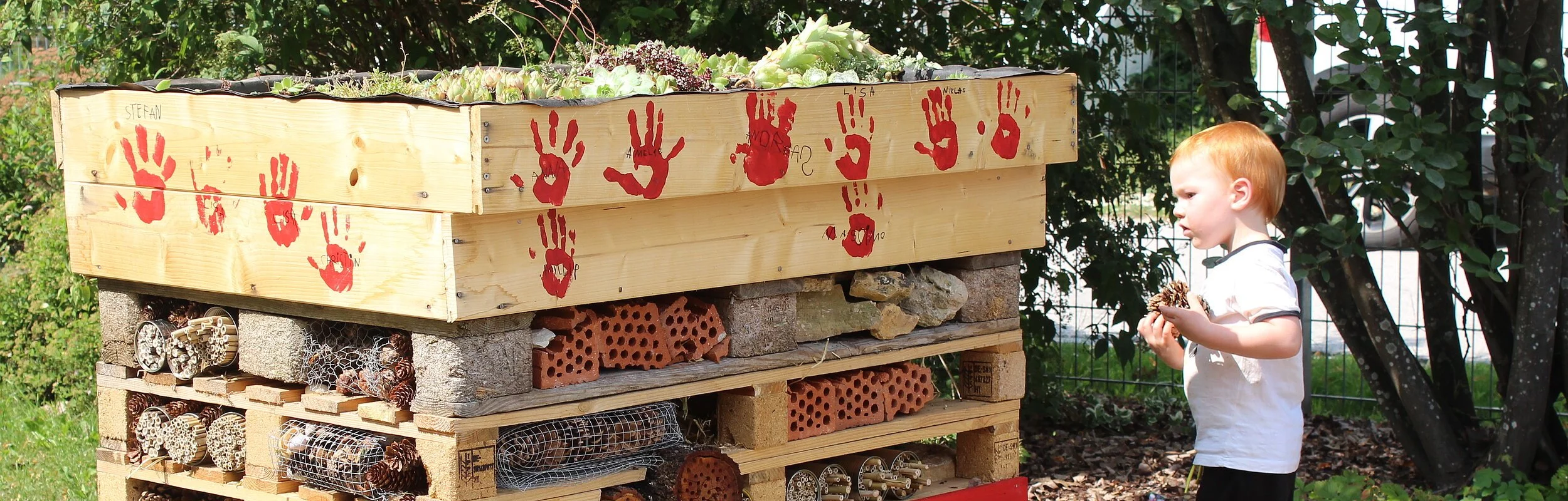 Ein kleiner Junge begutachtet das selbstgebaute Wildbienenhotel, auf dem die Handabdrücke der kleinen Helfer in rot zu sehen sind.