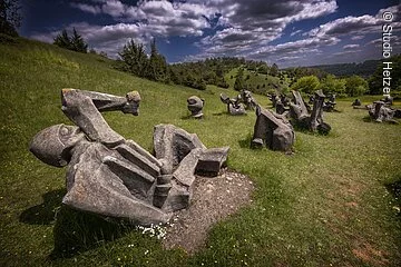Die Fotographie zeigt ein Skulpturenfeld, welches auf einer Wiese in Hanglage gelegen ist. Die abstrahierten, menschenartigen Figuren sind aus Beton gegossen und stellen Kämpfer und Opfer in verschiedenen Positionen dar. Die Gesamtheit des Figurenfeldes erinnert an ein Schlachtfeld. Der Hintergrund zeigt eine karge Berglandschaft.