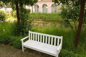 Die Aufnahme zeigt eine weiße Holzbank, die auf einem gepflasterten Weg steht und zu beiden Seiten von grünen Pflanzen umgeben ist. Dahinter befindet sich ein kleiner Teich mit schilfbewachsenem Ufer. Hinter diesem wiederum steht ein lachsfarbenes Gebäude. An dessen Fassade befinden sich drei große, bogenförmige Fenster. Um die Fenster zeichnen sich weiße Verzierungen ab. Ebenso sind vor den Fenstern weiße Gitter angebracht.