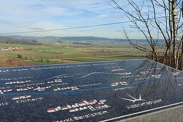 Im Vordergrund eine Steintafel. Dahinter eine weite grüne Landschaft. Felder und Bäume.