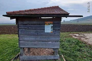 Kleine offene Holzhütte auf einer Wiese. Dahinter ein braunes Feld. Der Himmel ist bewölkt.