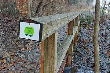 Eine Holzbrücke bedeckt mit Herbstlaub führt entlang von Bäumen und Sträuchern. Der Boden ist nass vom Regen. Am Brückengeländer Hämgt einSchild. Darauf eine grüne apfelgrafik mit der Aufschrift: Streuobst Erlebnislandschaft