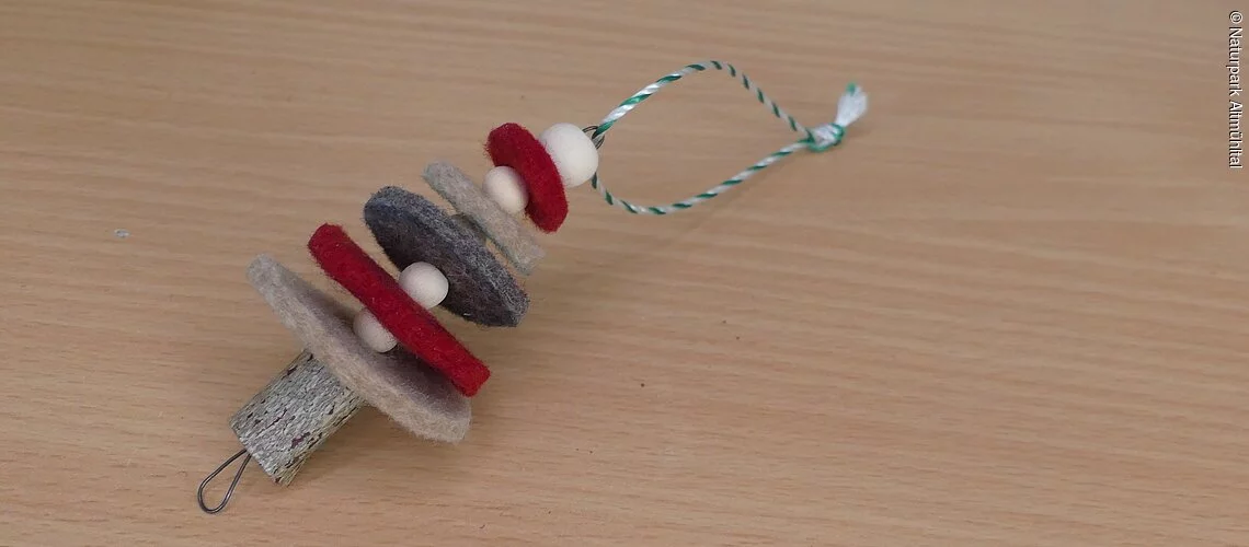 Ein Weihnachtsbaum-Anhänger aus rotem und grauem Filz liegt auf einem Holztisch