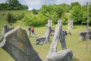 Auf den Wiesen im Hessental sind 78 überlebensgroße Plastiken, die zu einem fesselnden Mahnmal gegen Krieg und Gewalt geschaffen wurden, zu sehen. Inmitten dieses sind zwei Wanderer mit Rucksäcken unterwegs.