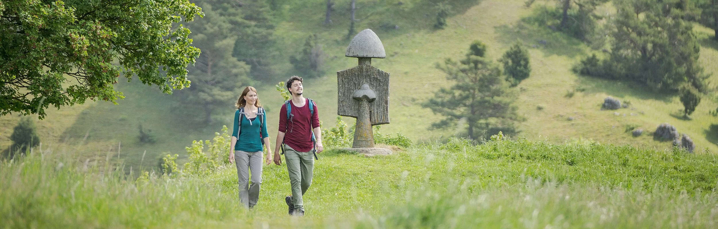 Ein Wanderpärchen wandert im Hessental. Sie sind umgeben von grünen Wiesen. Hinter ihnen ragt eine Figur des Figurenfeldes und Bäume am Hang entlang.