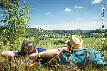 Wanderer an der Arnsberger Leite auf dem Altmühltal-Panoramaweg unterwegs Ein Mann mit verschränkten Armen und daneben eine Frau mit Strohhut, die ihren Kopf auf ihren blauen Rucksack abgelegt hat, liegen im Gras. Sie genießen den Blick von der Arnsberger Leite ins Tal. Der Himmel strahlt blau und nur ein paar Wolken haben sich dazu verirrt.