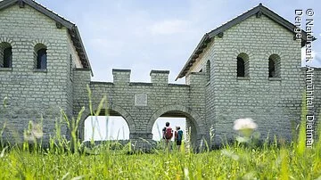 Römerkastell Vetoniana bei Pfünz Zwei Wanderer stehen am Nordtor des Römerkastells. Das Kastell wurde rekonstruiert und mit hellen Steinen gebaut. Rechts und links sind zwei Türme mit Dächern, die mit zwei Rundbögen miteinander verbunden sind.