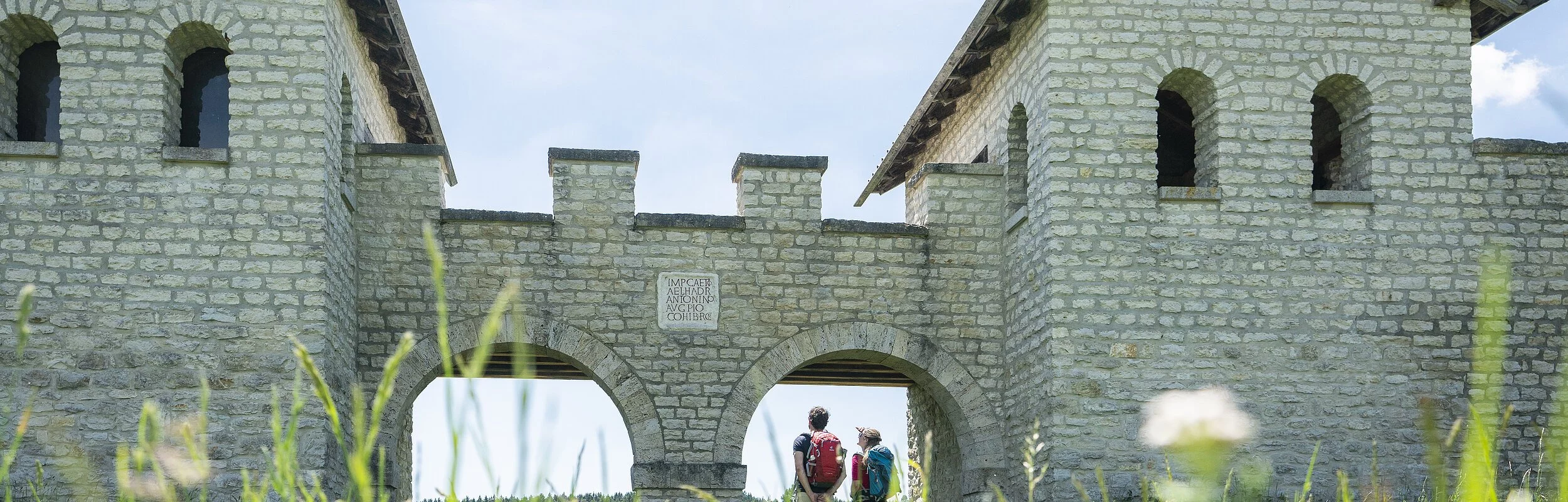 Zwei Wanderer stehen am Nordtor des Römerkastells. Das Kastell wurde rekonstruiert und mit hellen Steinen gebaut. Rechts und links sind zwei Türme mit Dächern, die mit zwei Rundbögen miteinander verbunden sind.