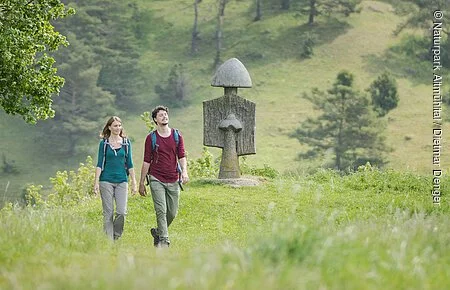 Ein Wanderpärchen wandert im Hessental. Sie sind umgeben von grünen Wiesen. Hinter ihnen ragt eine Figur des Figurenfeldes und Bäume am Hang entlang.