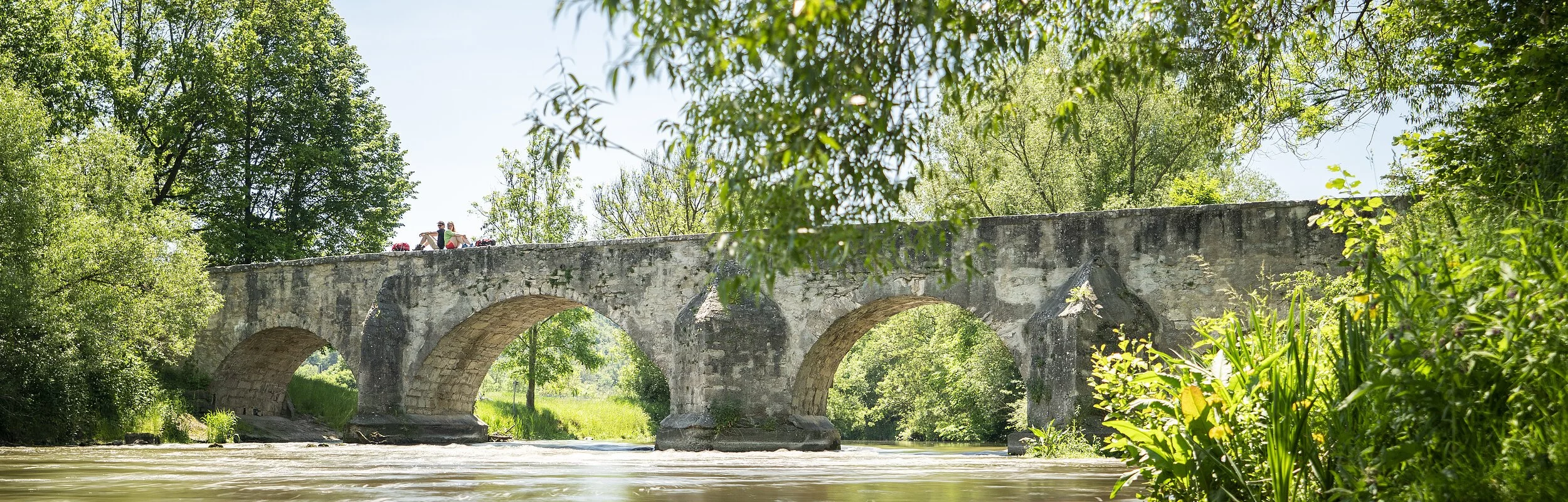 Auf der Römerbrücke sitzen zwei Wanderer Rücken an Rücken auf dem Steingeländer der Brücke. Eingerahmt von Bäumen und unter ihnen die Altmühl.