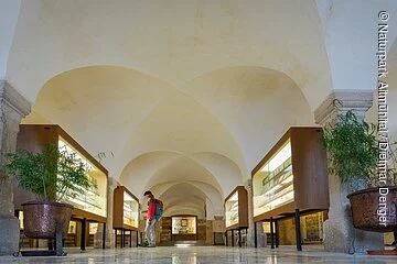 Im Mittelgang des Museums stehen links und rechts beleuchtete Vitrinen. Zwei Wanderer betrachten eine Vitrine auf der linken Seite. Der Raum ist hell und hat zudem Säulen und Rundbögen.
