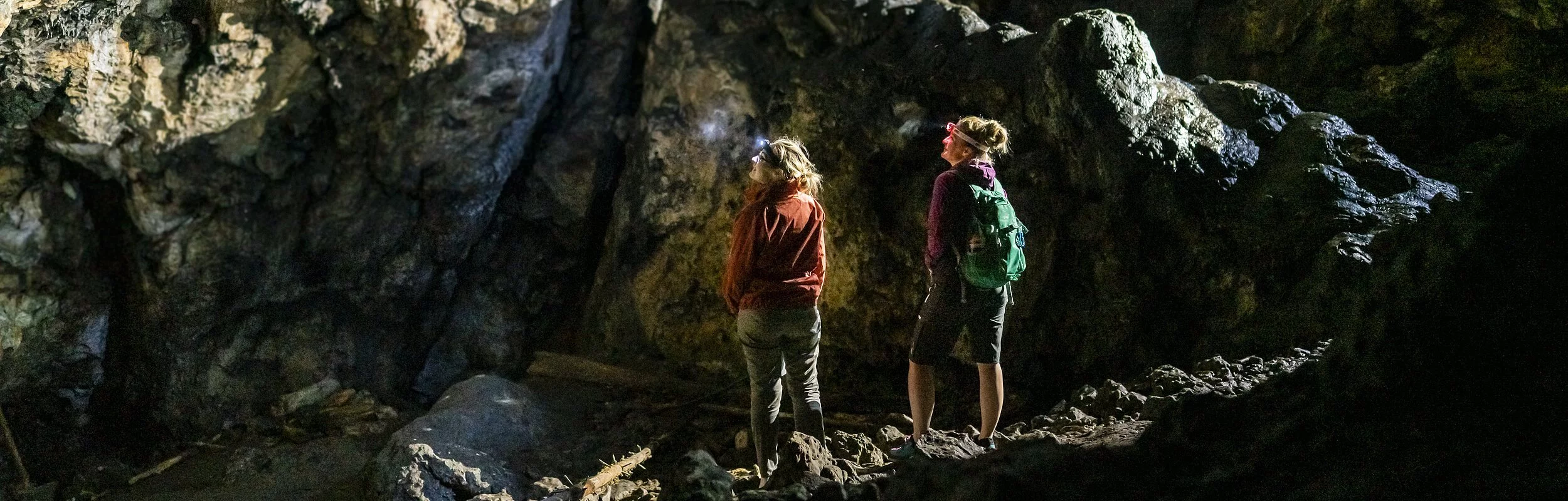 In der felsigen, dunklen Höhle, die über Treppenstufen zu erreichen ist. Stehen zwei Wanderer und schauen hoch zu einem Dritten, der über ihnen steht und mit einer Stirnlampe ausgerüstet ist.