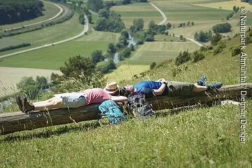 Auf der langen Bank liegen eine Frau links und ein Mann rechts. Ihre Rucksäcke an die Bank gelehnt. Hinter den beiden hat man einen guten Blick ins Tal.
