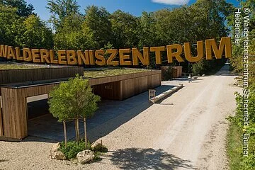 Walderlebniszentrum Schernfeld Am Walderlebniszentrum wird man am Eingang mit dem großen Schriftzug aus Holz empfangen. Darunter sieht man eine Holzhütte und rechts geht ein Feldweg in den Wald.