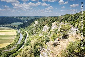 Sommerlicher Blick ins Altmühltal auf das Tal, auf den Fluss und auf die Felsenkette Arnsberger Leite, auf der ganz klein am Talrand eine Familile steht und ins Tal schaut.