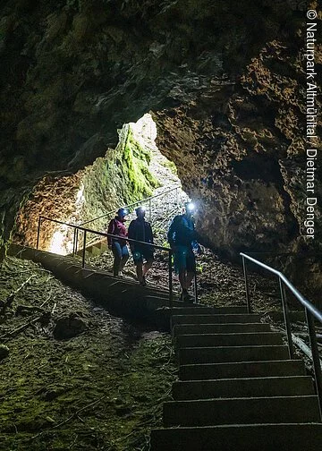 Arndthöhle bei Attenzell Drei Wanderer steigen die Treppenstufen zur Höhle hinab. Der Vorderste ist mit einer Stirnlampe ausgestattet und leuchtet ihnen den Weg aus. Licht kommt nur vom Eingang in die Höhle.