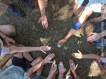 Kinder sitzen im Kreis und strecken ihre Hände in die Mitte. Sie halten jeweils ein keltisches Amulett in der Hand.