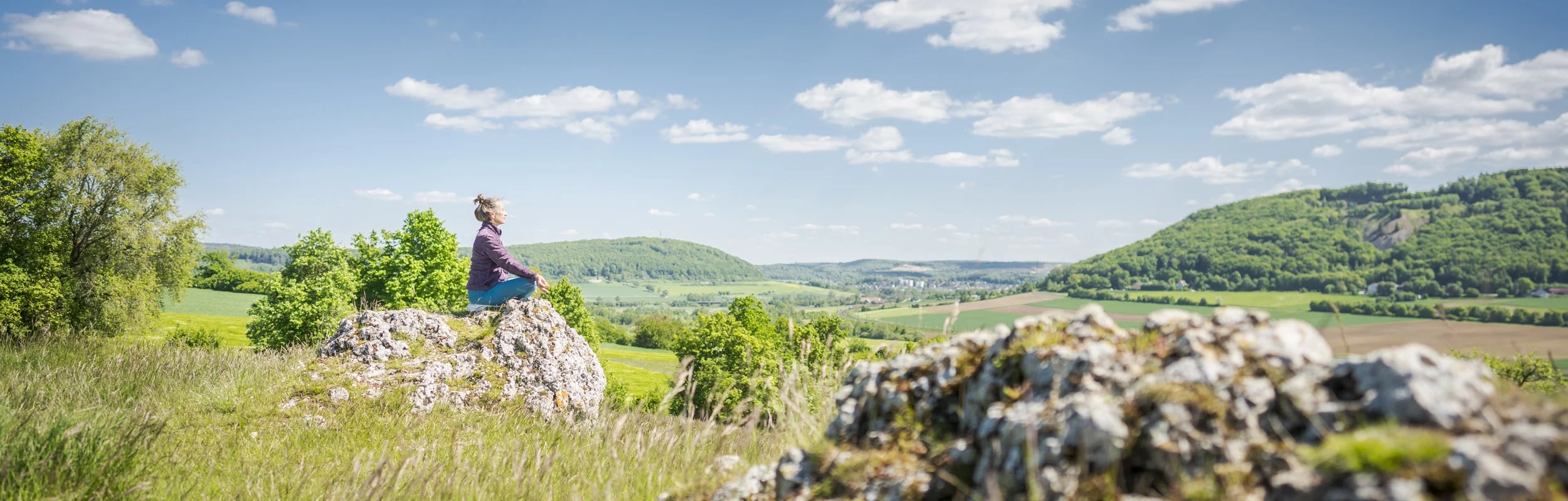 Frau sitzt auf einem Stein in Yogapose im Vordergrund ein Felsen