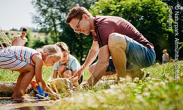Wasserspielplatz Enkering Ein Vater spielt mit zwei Kinder – einem Mädchen und einem Jungen – in einem flachen Bachlauf.