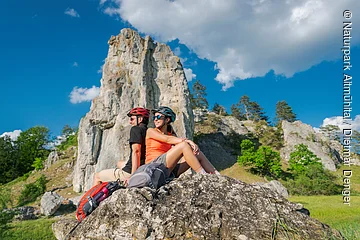 Ein Mann und eine Frau mit Fahrradhelm sitzen auf einem Felsen vor dem Burgsteinfelsen und machen Rast.