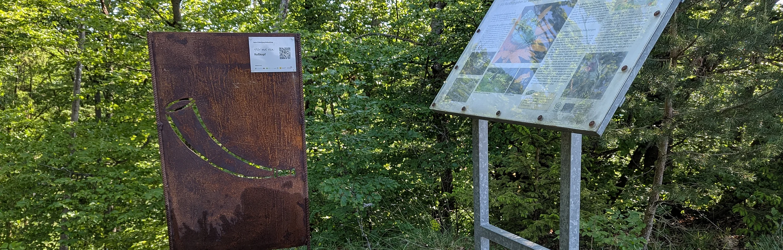 braune Metallstele und weiße Infotafel vor grünen Bäumen