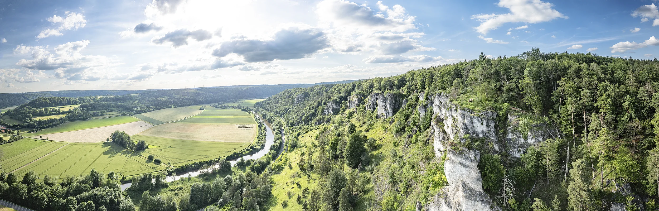 Weiter Blick ins Altmühltal: Rechts Felsen mit grünem Hang ins Tal  und rechts sieht man den Fluss im Tal mit Wiesen.