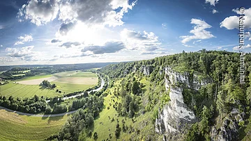 Weiter Blick ins Altmühltal: Rechts Felsen mit grünem Hang ins Tal  und rechts sieht man den Fluss im Tal mit Wiesen.