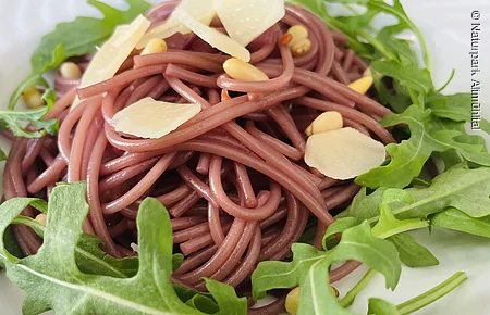 Rote Spaghetti mit Rucola, Pinienkernen und Käsespänen auf einem weißen Teller.