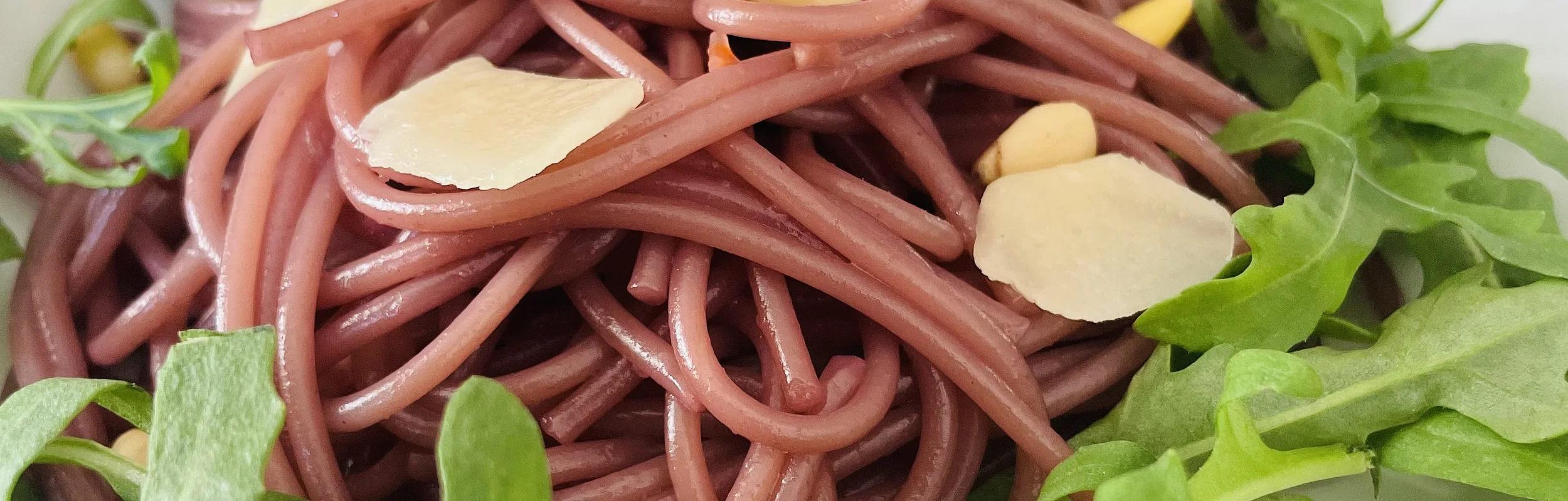 Rote Spaghetti mit Rucola, Pinienkernen und Käsespänen auf einem weißen Teller.