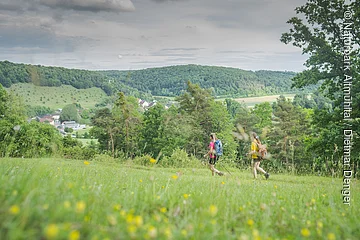 Zwei Personen wandern über eine Wiese mit Blick auf ein Dorf und bewaldete Hügel im Hintergrund.
