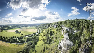 Arnsberger Leite mit Blick ins Altmühltal Landschaft mit grünen Feldern, einem Fluss und bewaldeten Hügeln unter einem bewölkten Himmel.