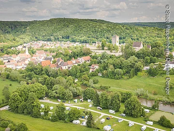 Luftaufnahme eines Dorfes mit Kirche, Burg und Wohnmobilen auf einem Campingplatz am Fluss.